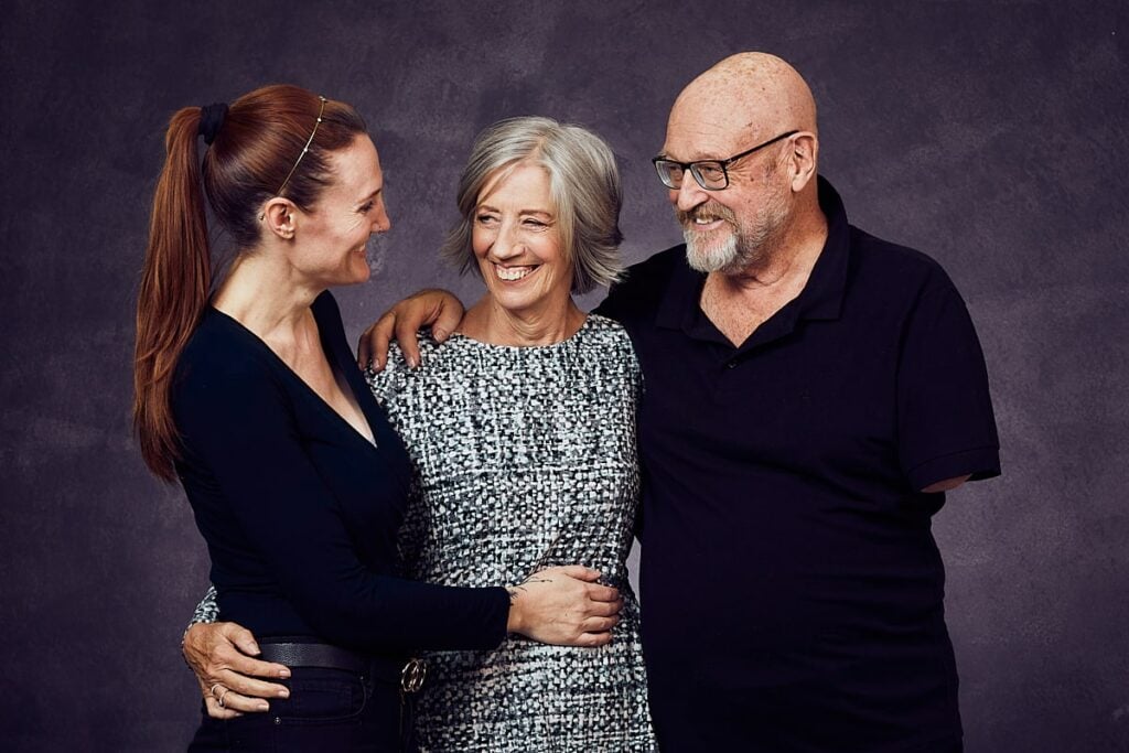 Lifestyle photography of a woman and her husband in their 60's with their daughter in her 30's. The woman has grey hair and is dressed in a white, knitted dress and the man and the daughter are dressed in all black. Shot in studio in Cape Town, South Africa